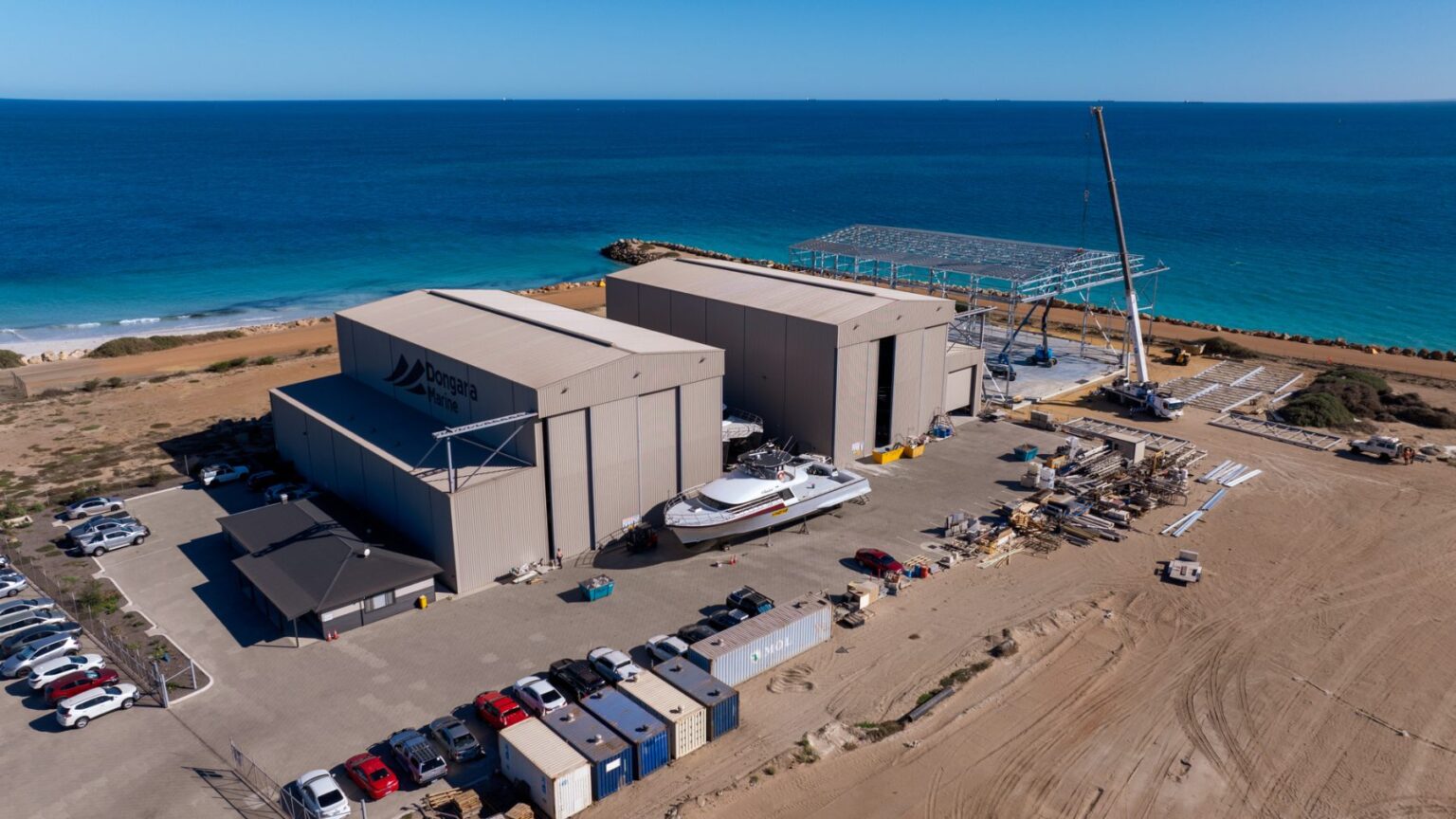 Aerial view of the Dongara Marine boat building workshops on the coast, featuring large warehouses, a moored boat, shipping containers, and a construction crane.