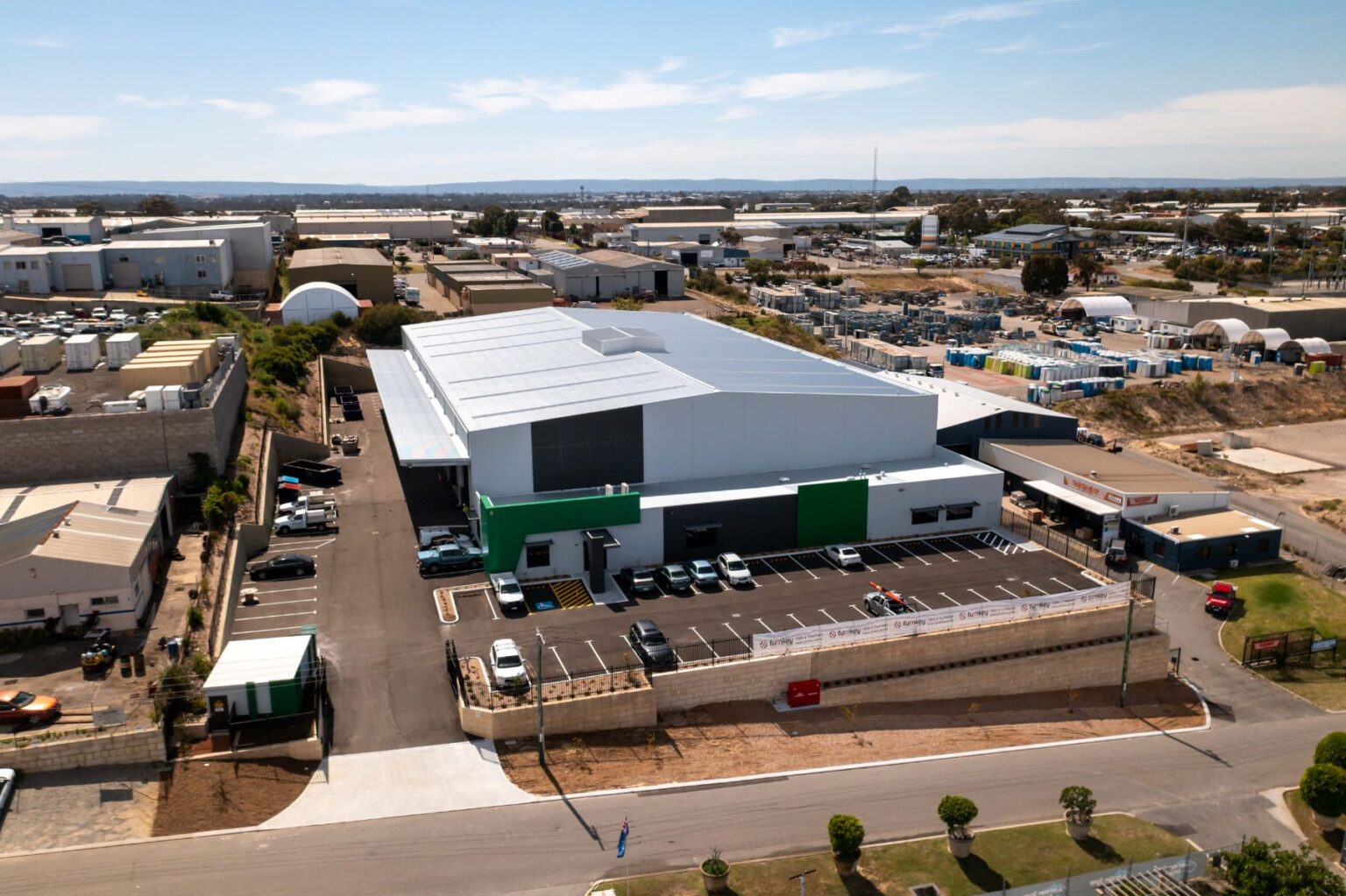 Aerial view of a large industrial factory building with an attached car park and surrounding commercial area.